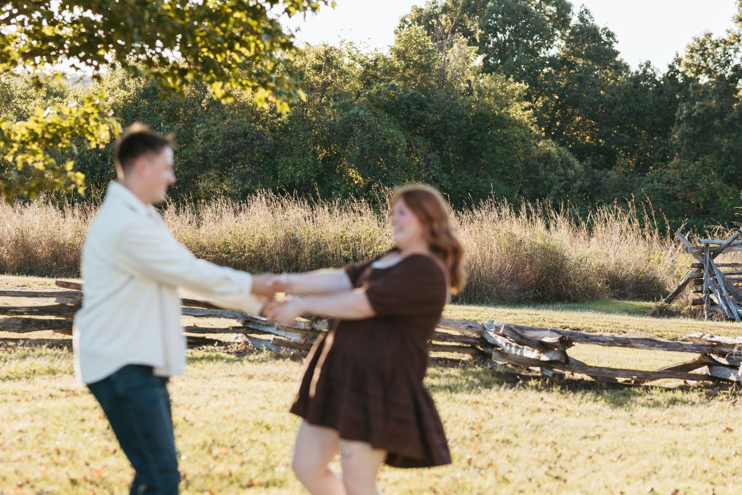A couple spins and dances together in a field during sunset with trees in the background.