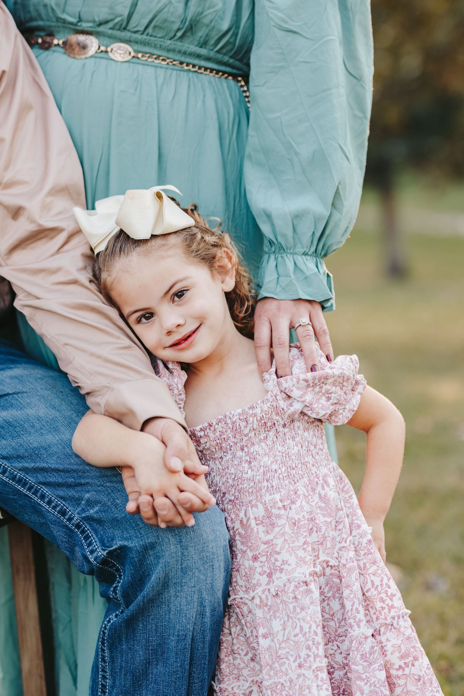 A child in a pink dress poses with parents wearing teal and denim in an outdoor setting.