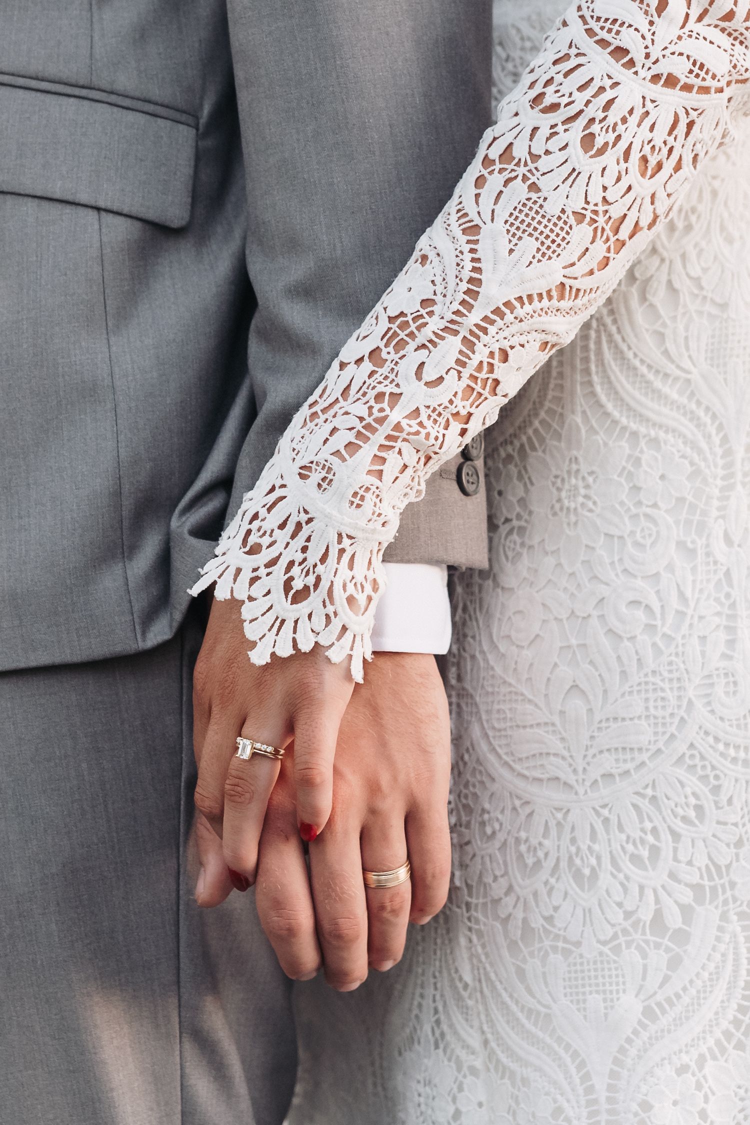 Close-up of intricately detailed white lace wedding dress sleeve against a gray suit with rings visible.