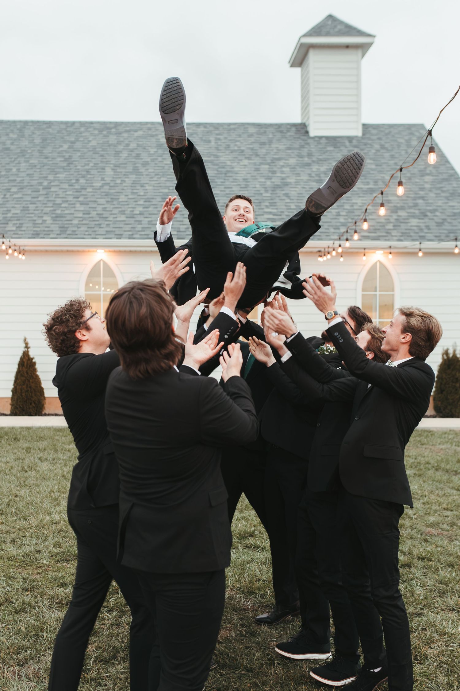 Groomsmen toss someone in suit into air during fun wedding photo outside white chapel.