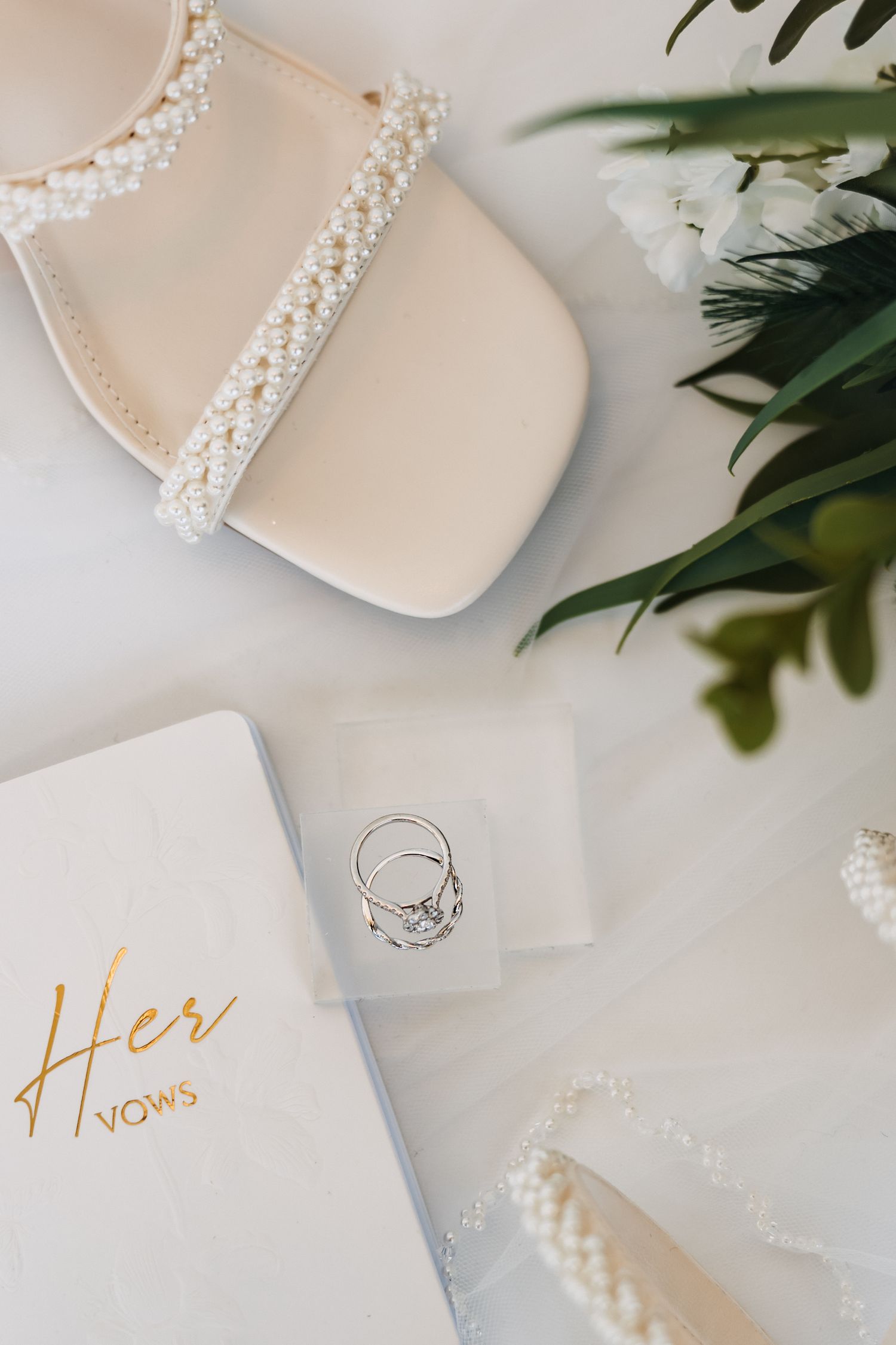 Wedding details including pearl-adorned shoes, vow book with gold lettering, and diamond ring.