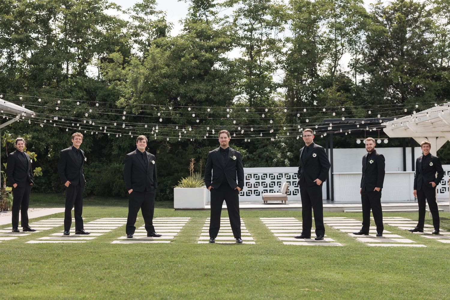 Wedding party members in black suits pose spaced apart on geometric outdoor patio.