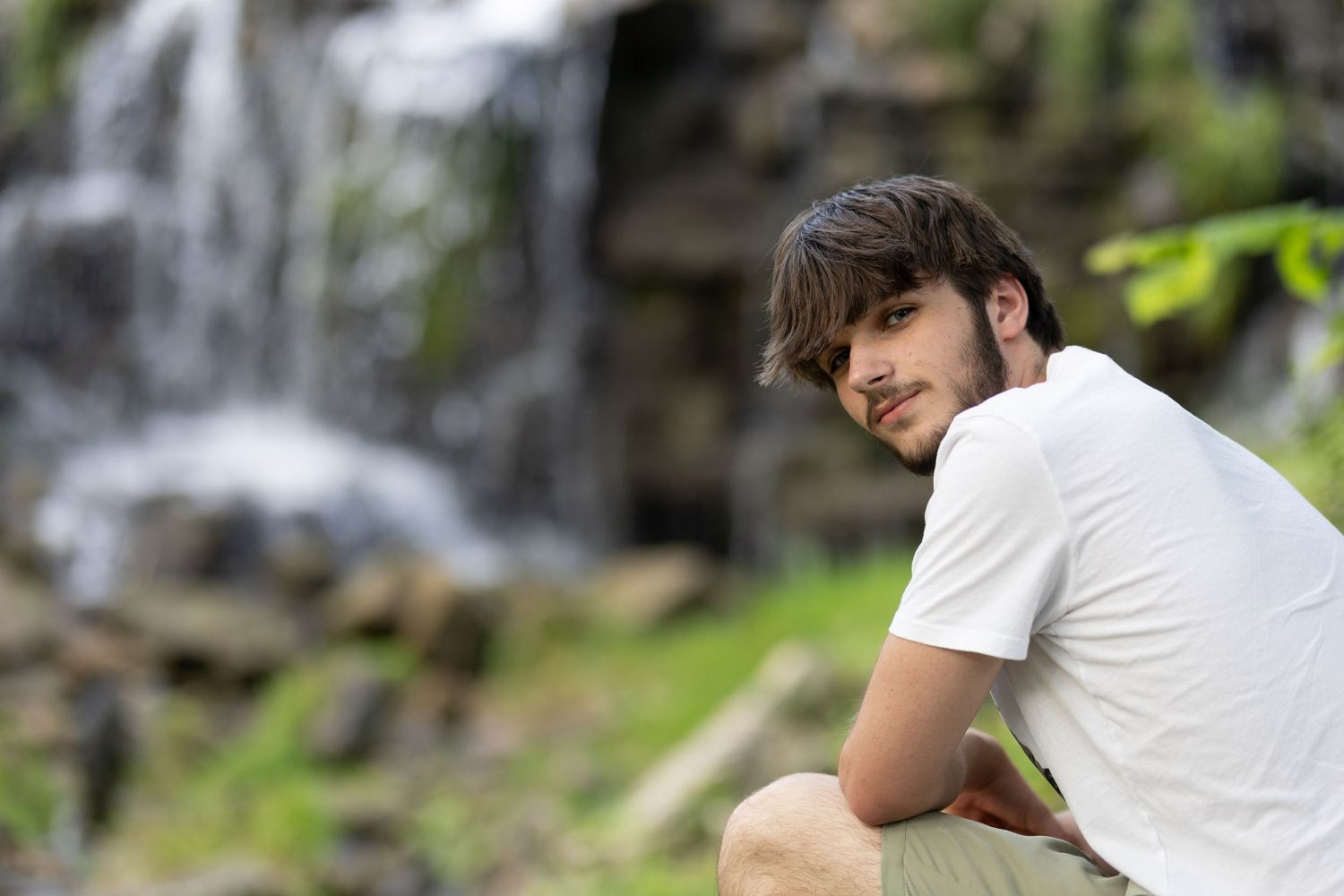 Person in white t-shirt sitting near a waterfall in nature setting.
