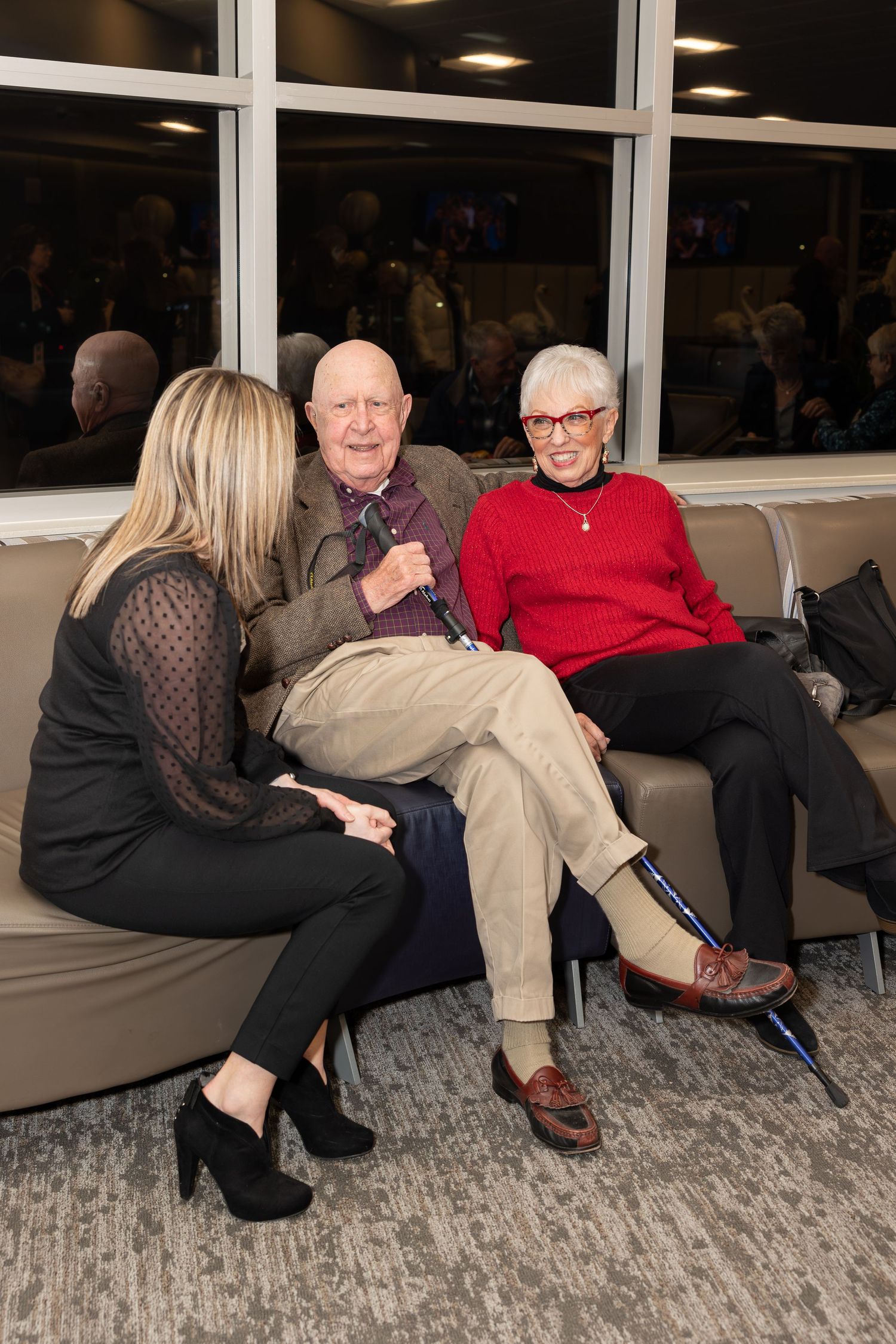 People having a conversation while seated on a gray couch in front of large windows.