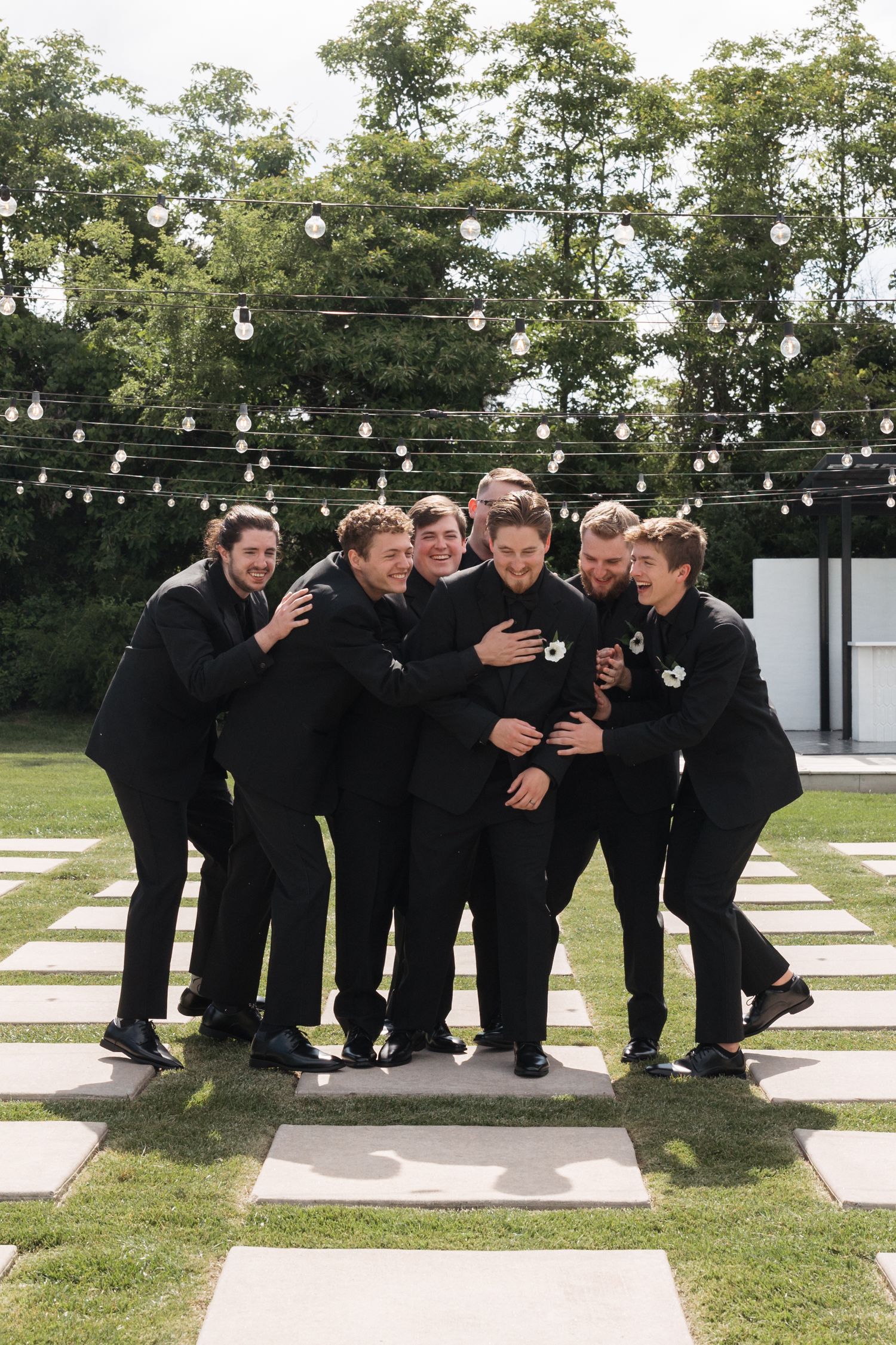 A group in formal black attire hugs and laughs together outdoors under string lights on a checkered pathway.
