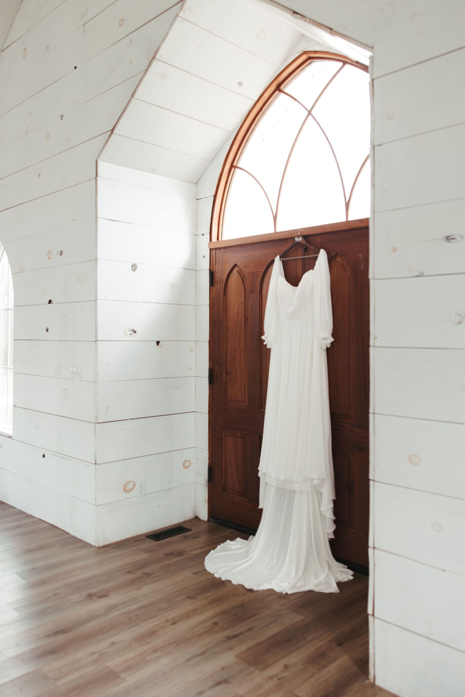 Vintage-style wedding dress hanging in white shiplap room with arched wooden door frame.