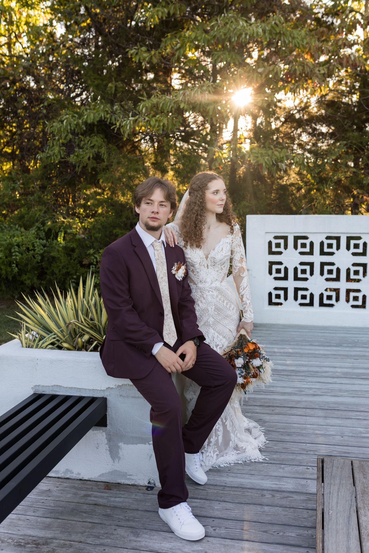 Bride and groom pose on a white bench at sunset, she in lace gown with bouquet, he in burgundy suit with white sneakers.