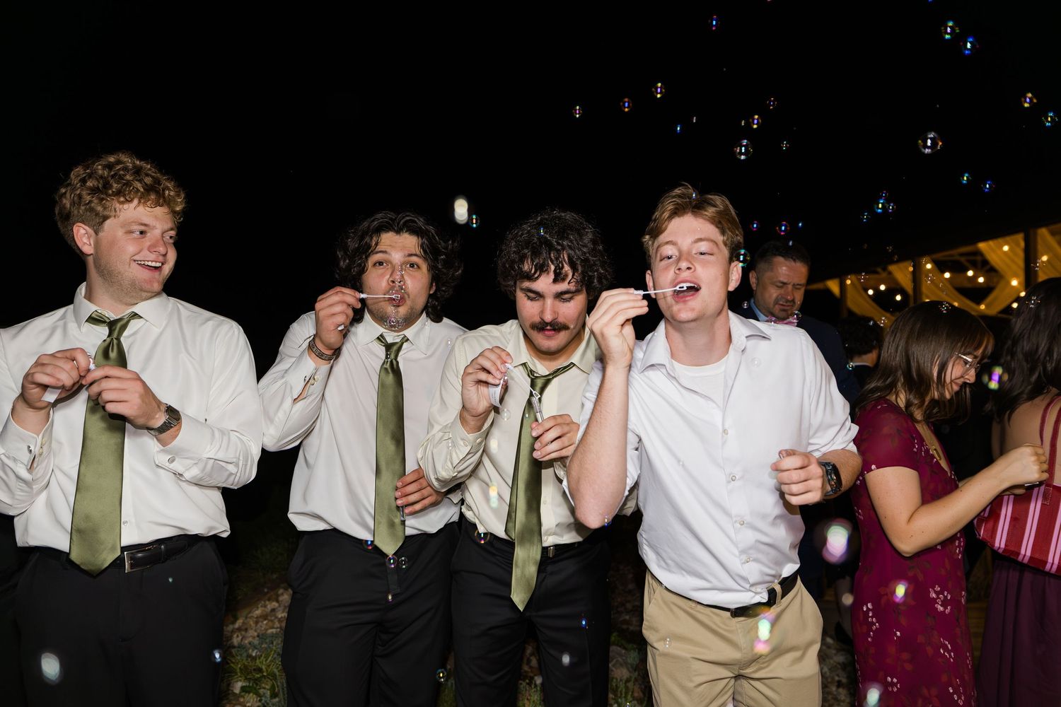 Five young men in white shirts and green ties laugh and blow bubbles at an outdoor evening wedding reception.