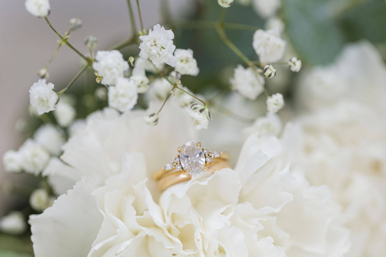 Gold solitaire diamond engagement ring resting on white hydrangea flowers with baby's breath in background.