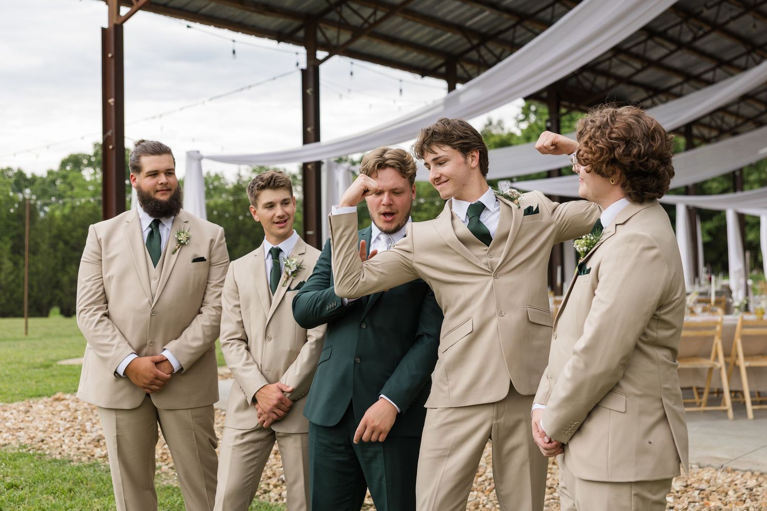 Groom in green suit laughs with groomsmen in tan suits at outdoor wedding venue.