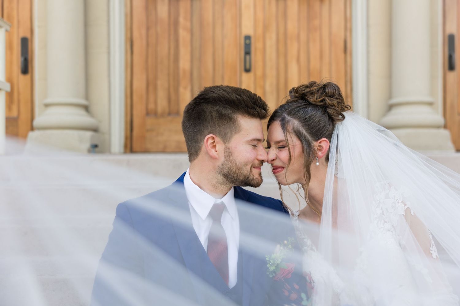 Bride and groom touching foreheads under flowing veil outside wooden church doors on wedding day.