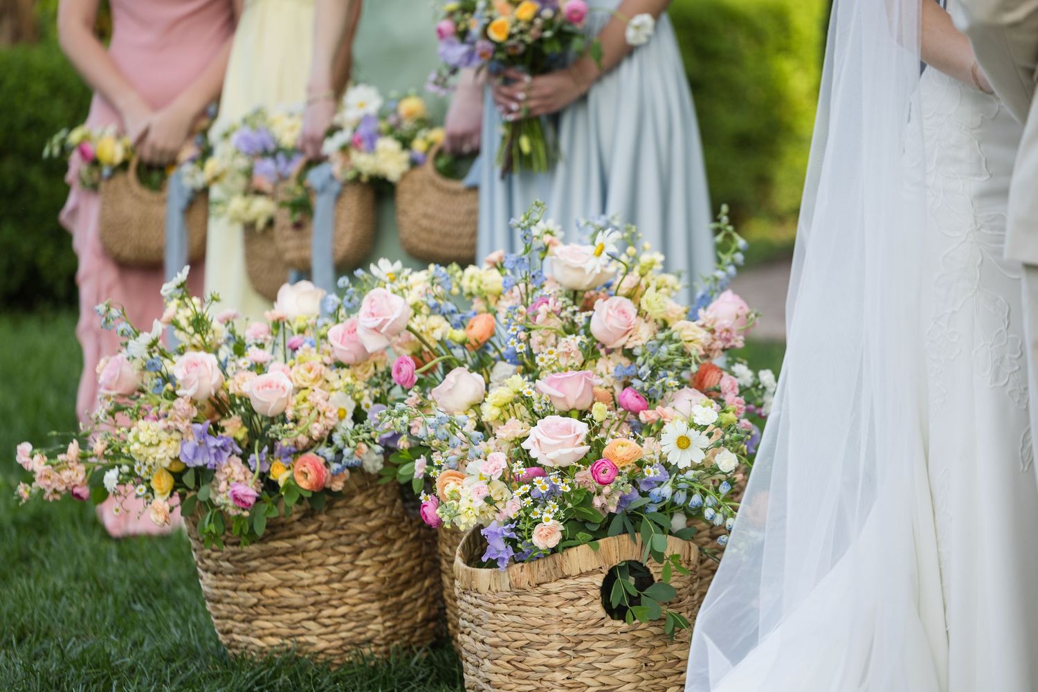 Bridesmaids holding wicker baskets overflowing with colorful spring flowers at an outdoor wedding ceremony.