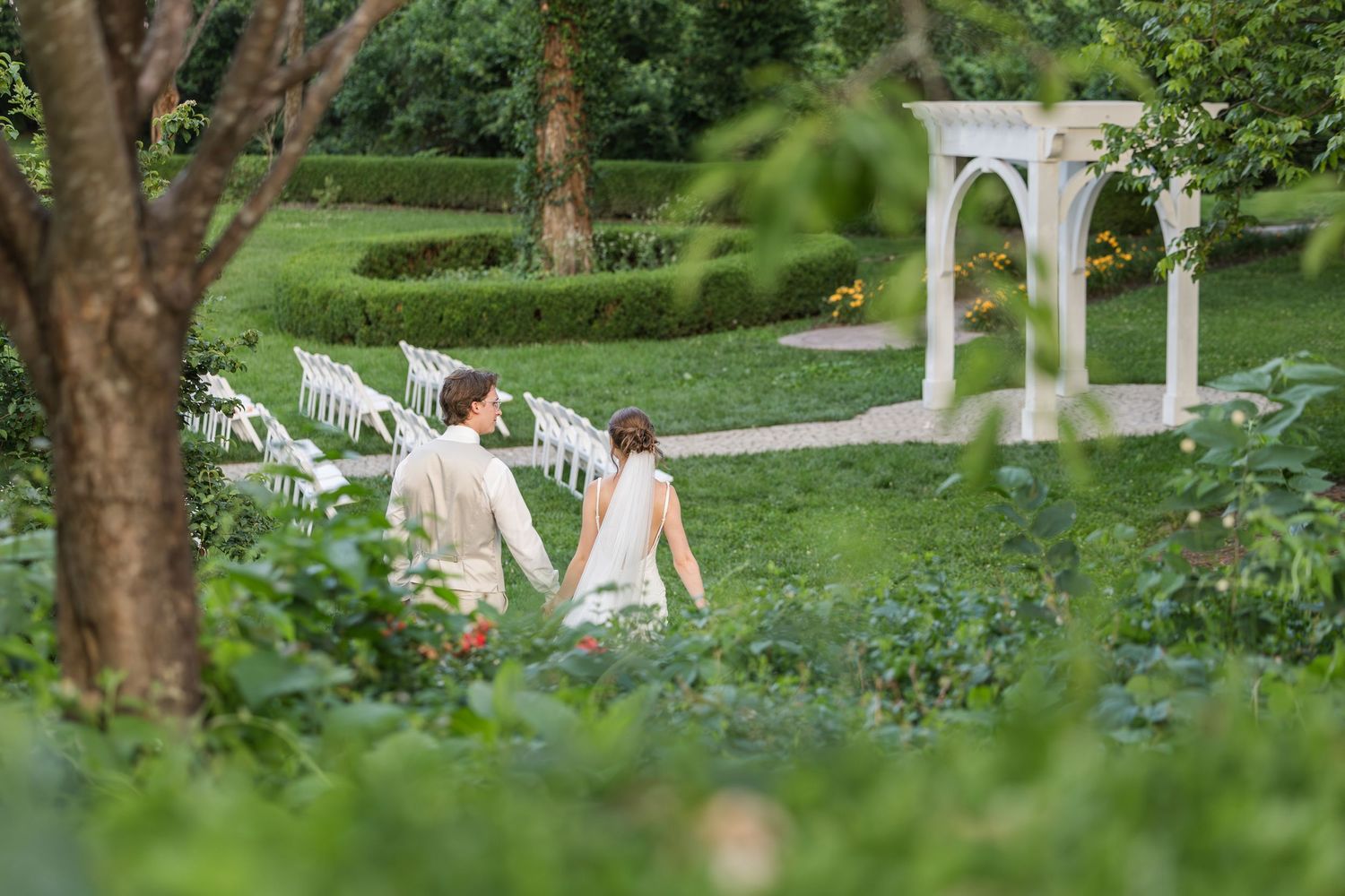 A couple walks hand-in-hand through a lush garden wedding venue with white chairs and elegant arch.