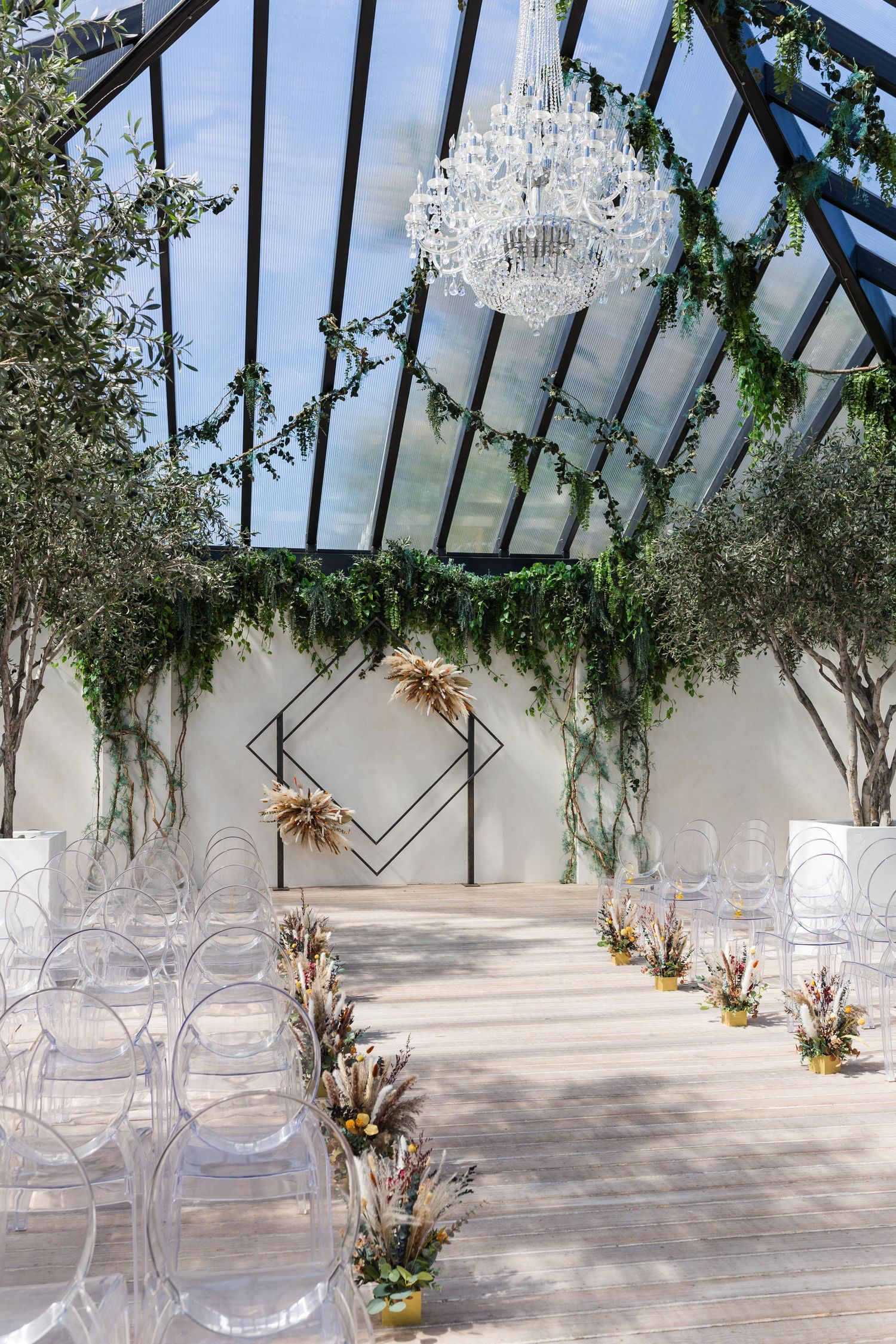 Elegant greenhouse wedding ceremony with crystal chandelier, ghost chairs, geometric arch, and lush greenery decor.