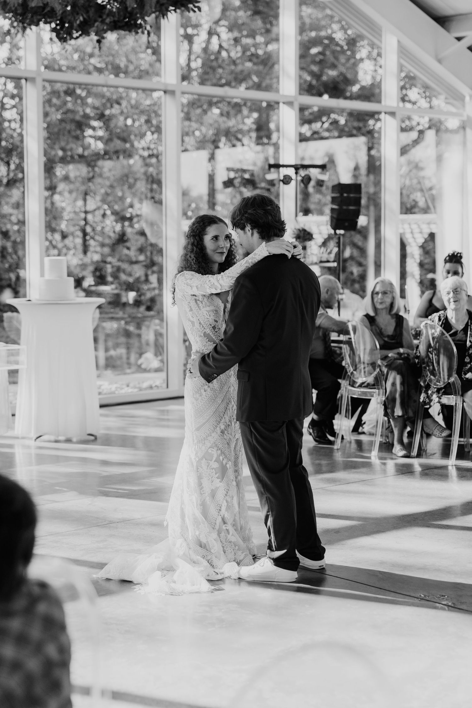 Black and white photo of bride and groom sharing first dance in elegant glass-walled reception venue.