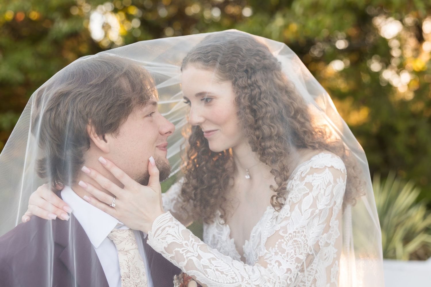 Bride and groom share an intimate moment together under a flowing white wedding veil outdoors.