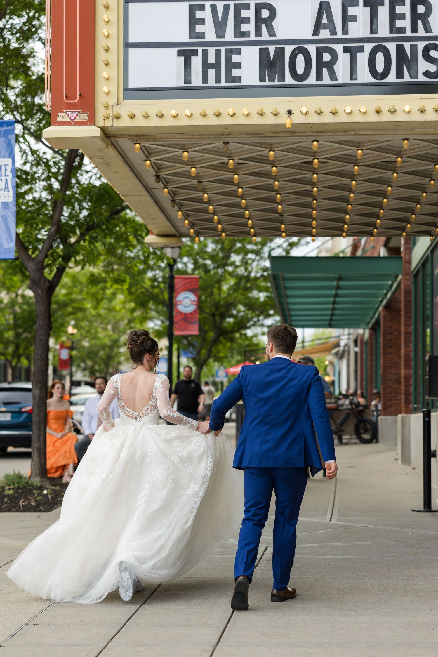 Bride and groom walk hand in hand under a theater marquee reading Ever After The Mortons.