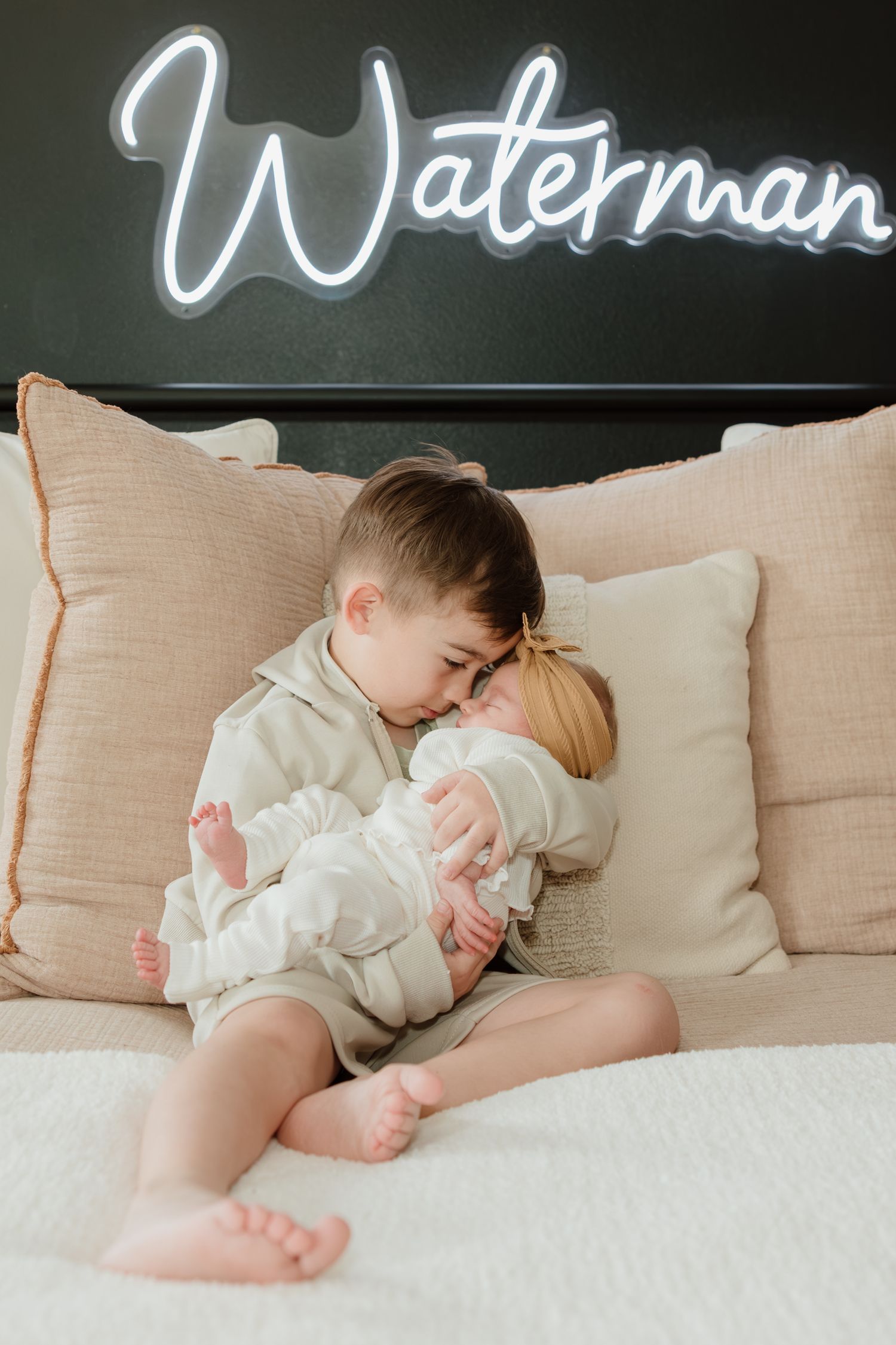 A heartwarming moment on a bed beneath a neon 'Waterman' sign shows two young siblings sharing a tender embrace.