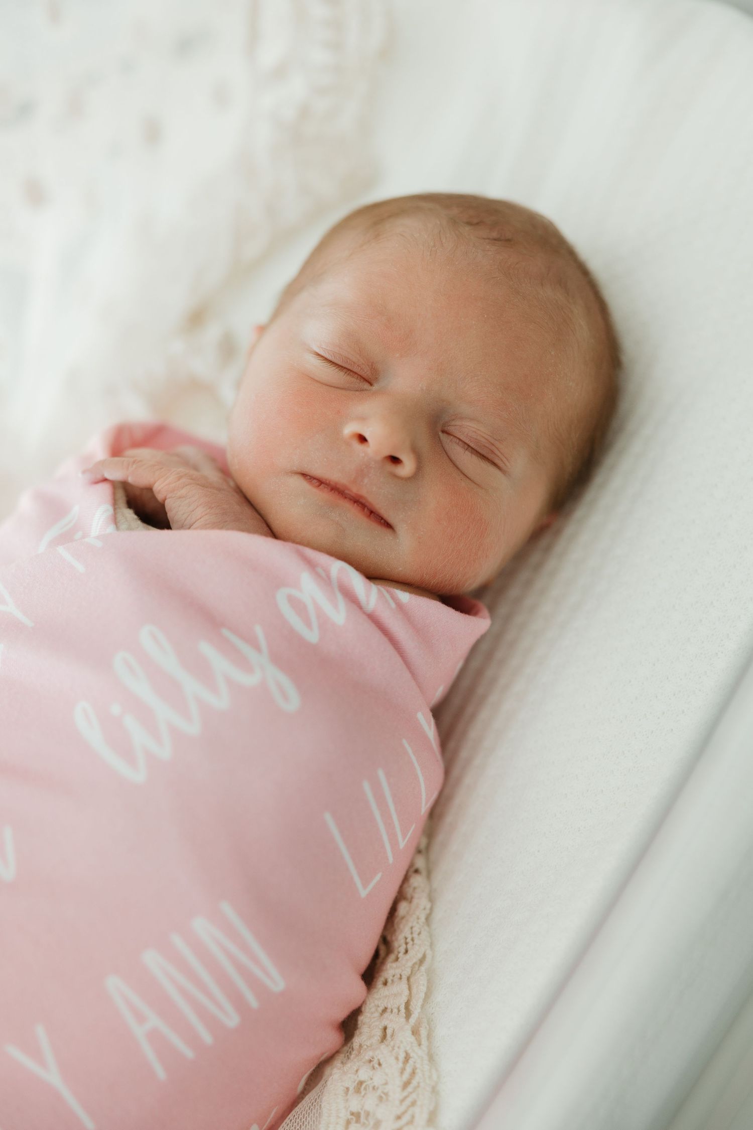 A sleeping newborn baby wrapped in a soft pink blanket with white text, resting peacefully on white bedding.