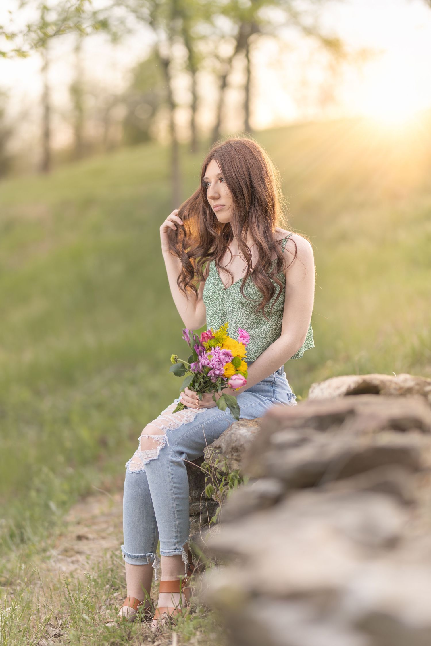 A woman in distressed jeans sits on rocks holding wildflowers at sunset in a peaceful meadow setting.