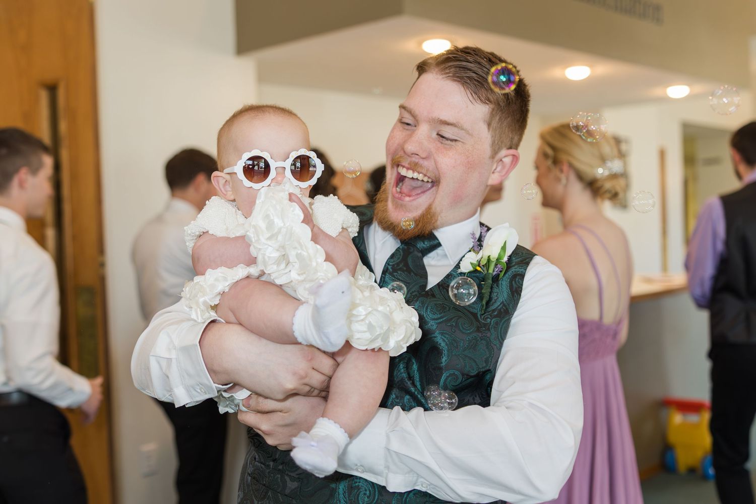 At a wedding reception, a joyful person in a vest holds a baby wearing white sunglasses and a lace dress.