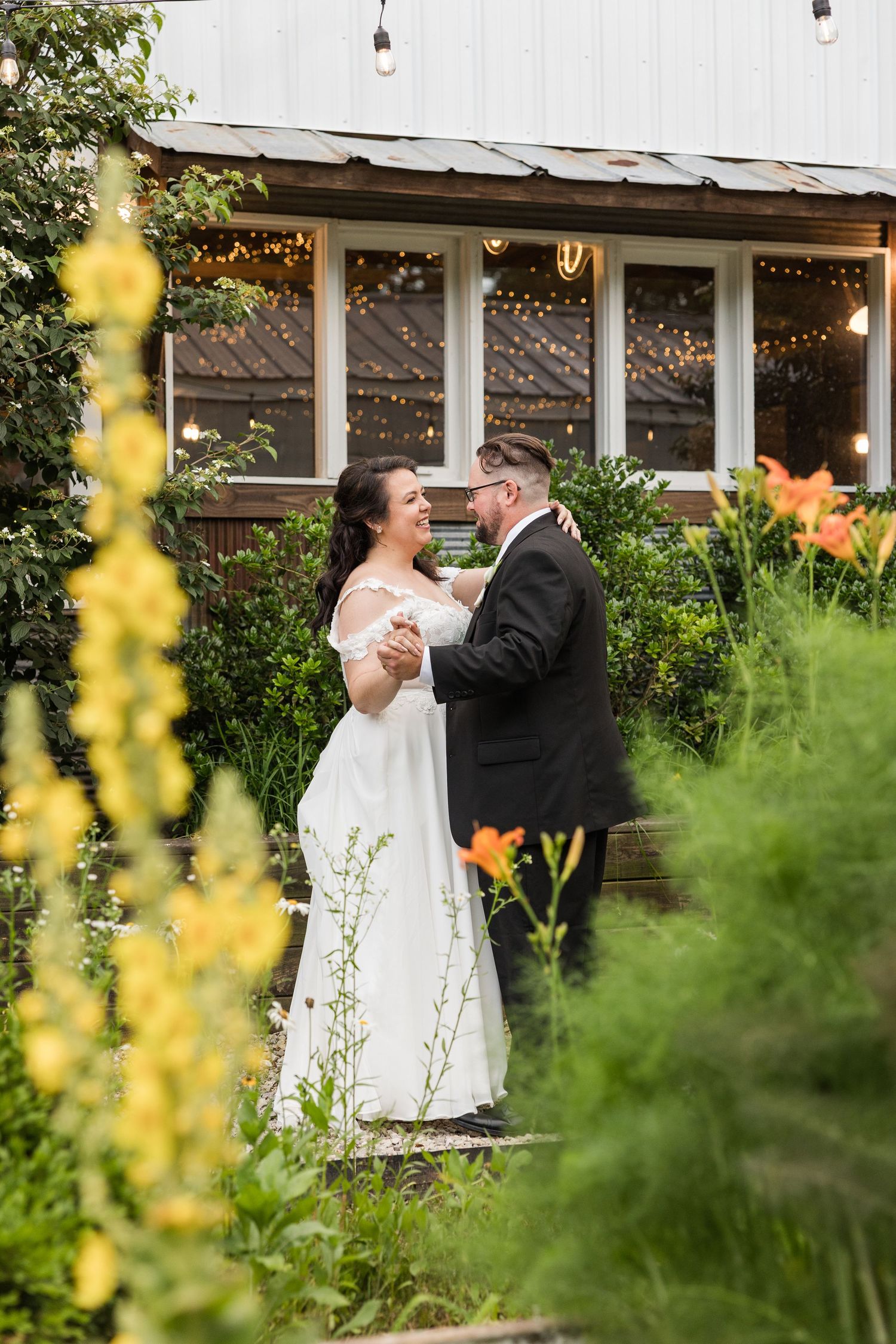 A romantic wedding couple shares a dance moment in a garden with yellow flowers and greenery in the foreground.