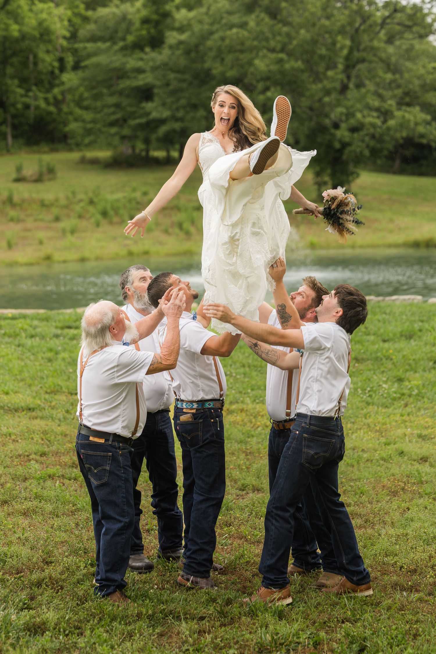 Groomsmen tossing bride in the air outdoors near a pond, wearing white shirts and suspenders at a rustic wedding.