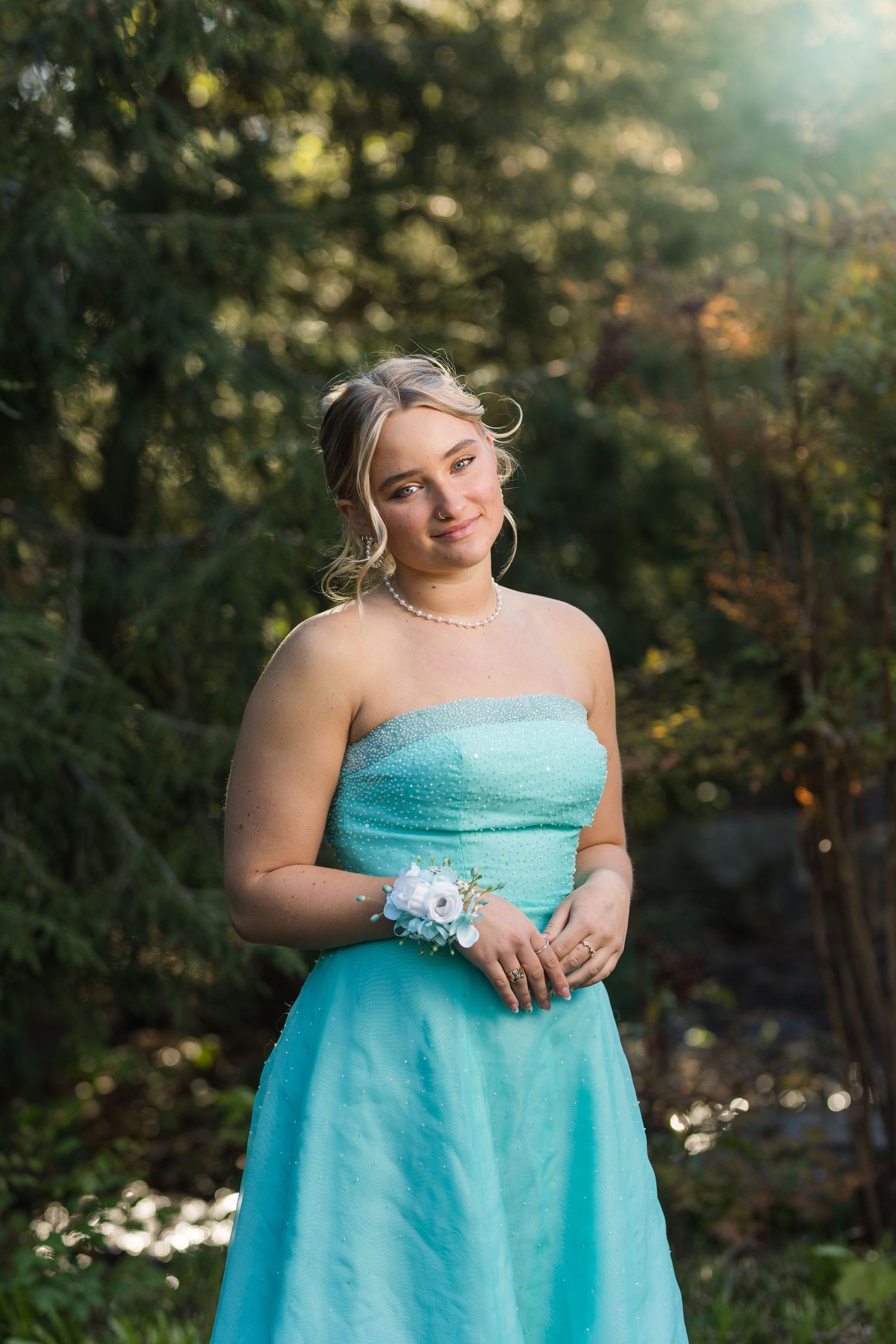 Young woman in a strapless teal prom dress with a floral wrist corsage and pearl necklace posing outdoors.