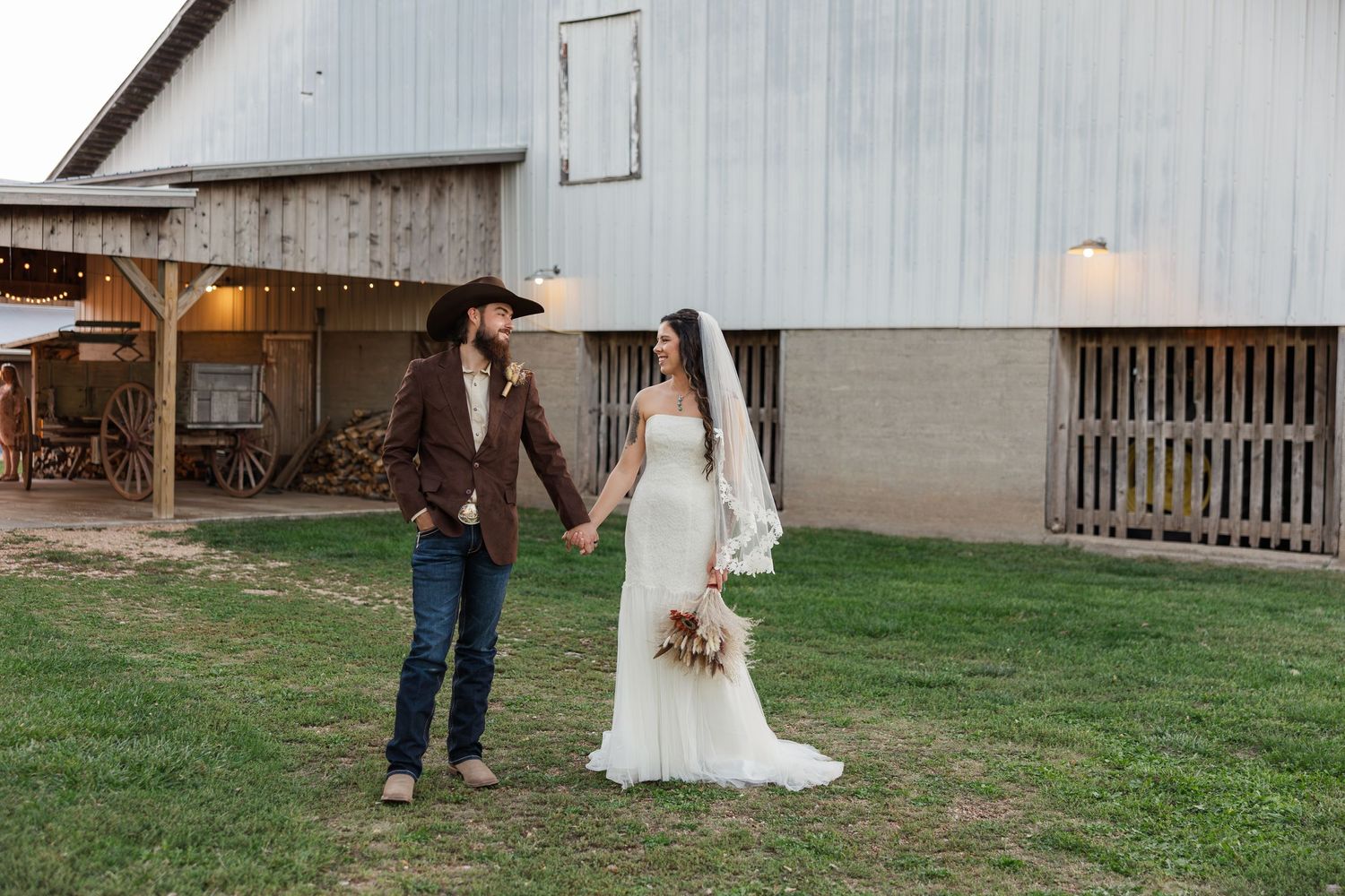 Bride and groom holding hands outside a rustic barn venue, groom in cowboy hat and brown jacket, bride in white gown.