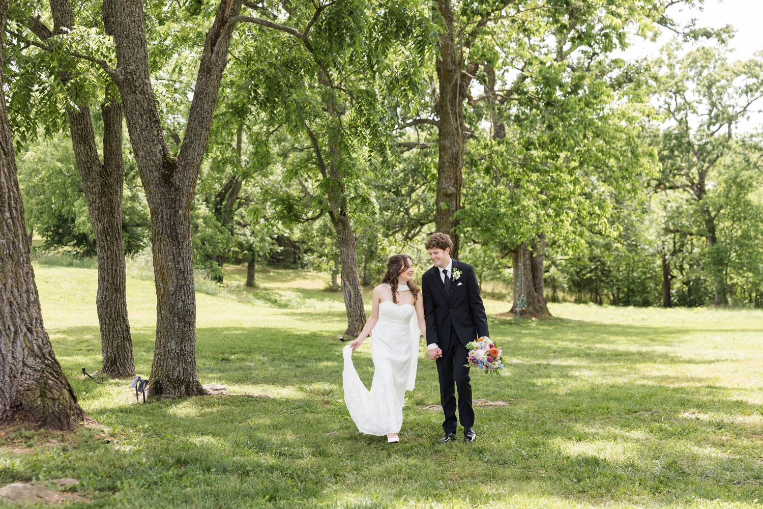 A bride and groom walk hand-in-hand through a lush green park surrounded by tall trees on their wedding day.