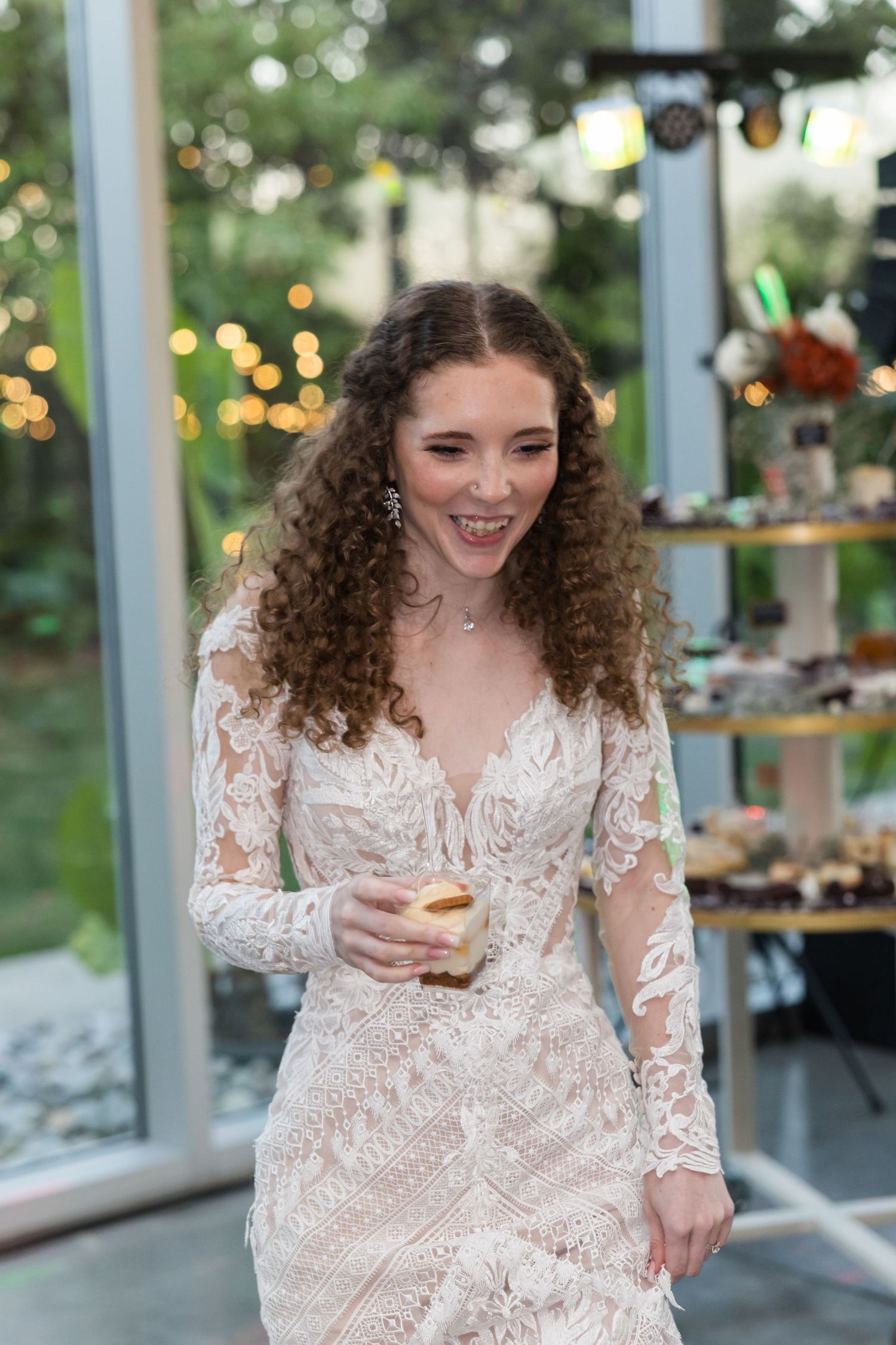 Joyful curly-haired bride in long-sleeve lace gown laughs while holding a drink at reception.