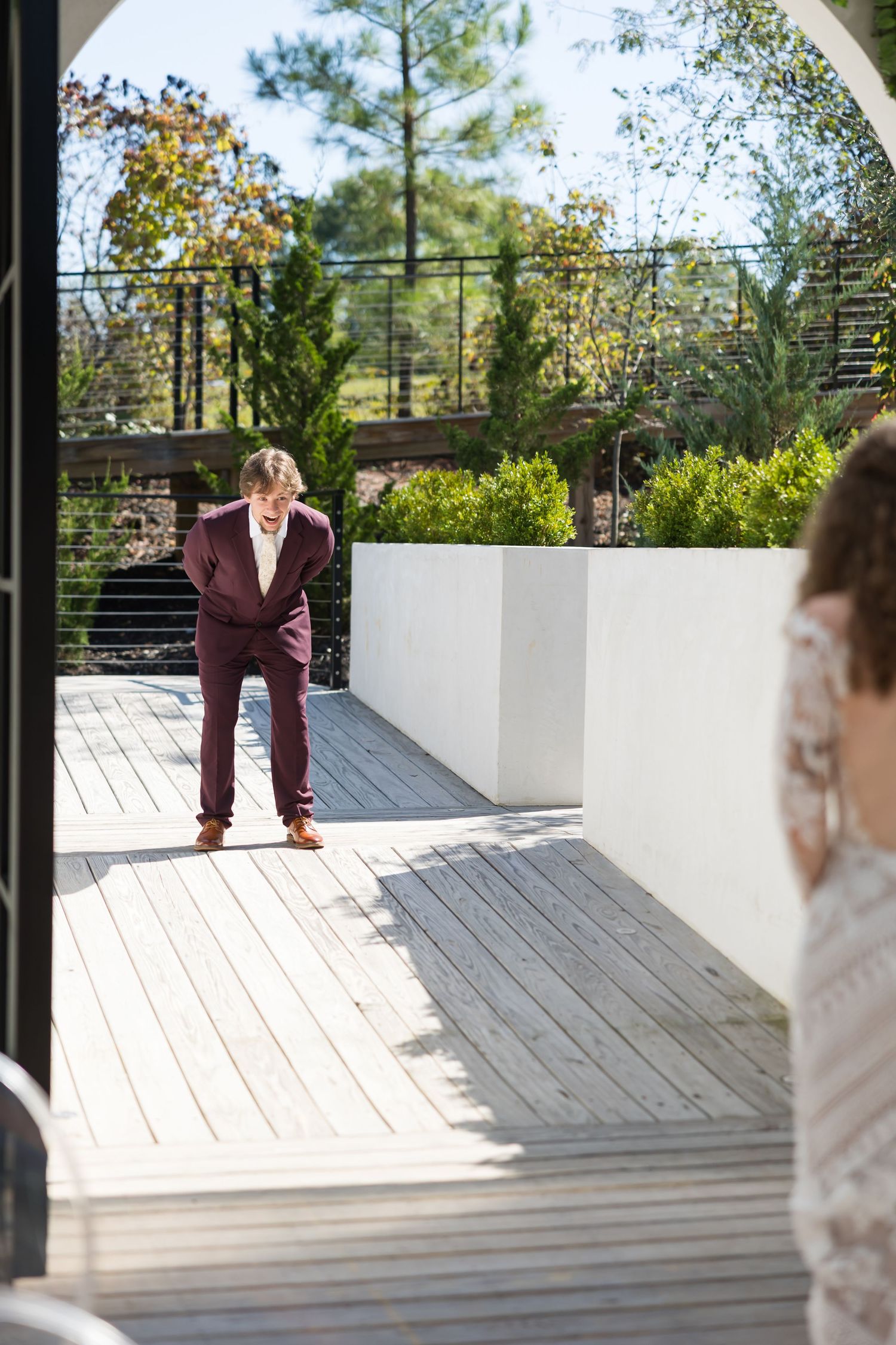 Groom in burgundy suit bowing on sunny outdoor deck as bride approaches in lace wedding dress.