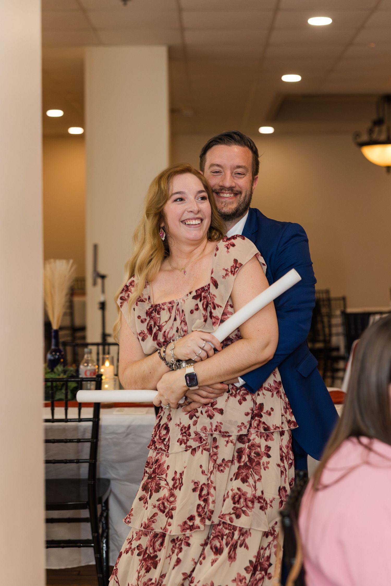 Smiling couple embraces at a wedding reception, woman in floral dress holding a rolled program scroll.