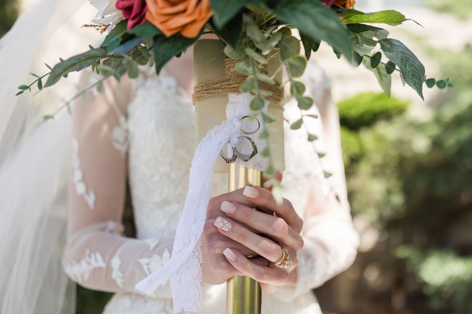 Bride in lace wedding dress holding a colorful bouquet with wedding rings draped over it.