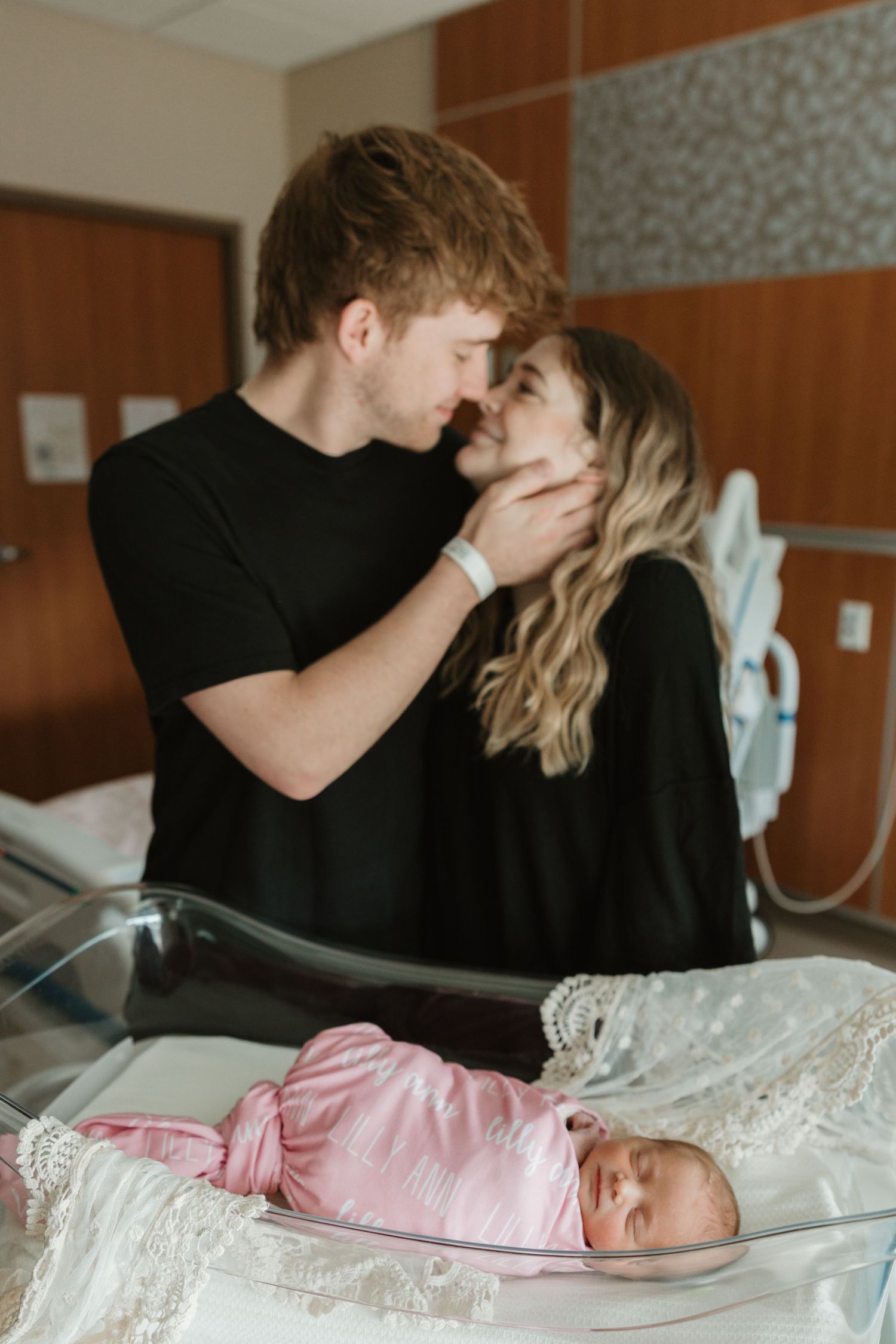New parents share an intimate moment above their newborn baby dressed in pink swaddled in a hospital bassinet.