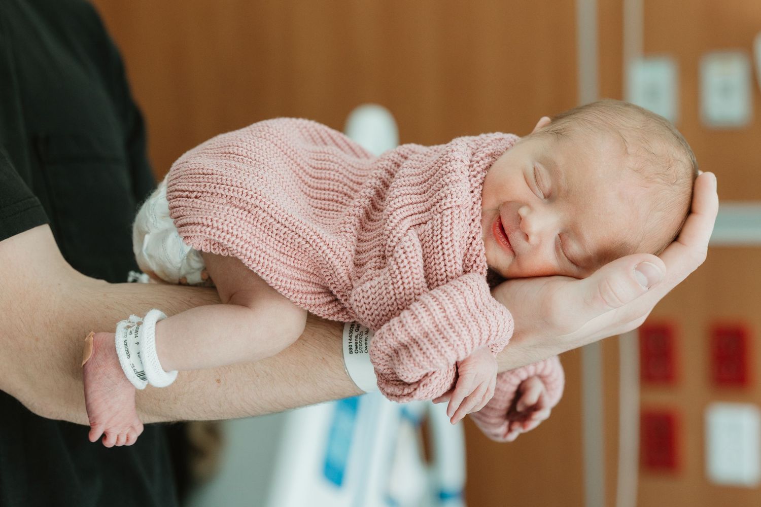 A newborn baby wearing a pink knitted sweater sleeps peacefully while being cradled in hands.