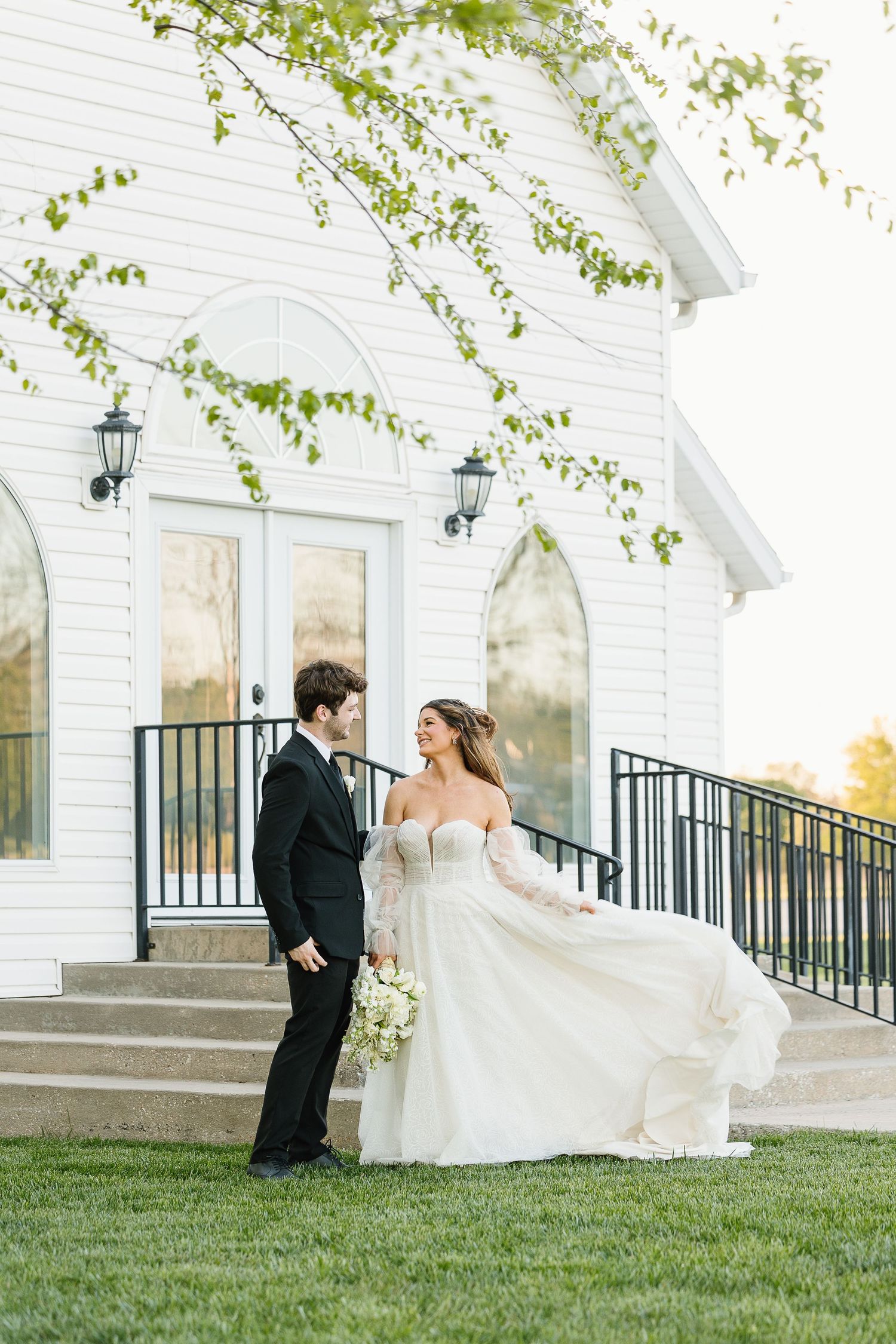A couple shares a romantic moment outside a white chapel with spring trees and black railings in the background.