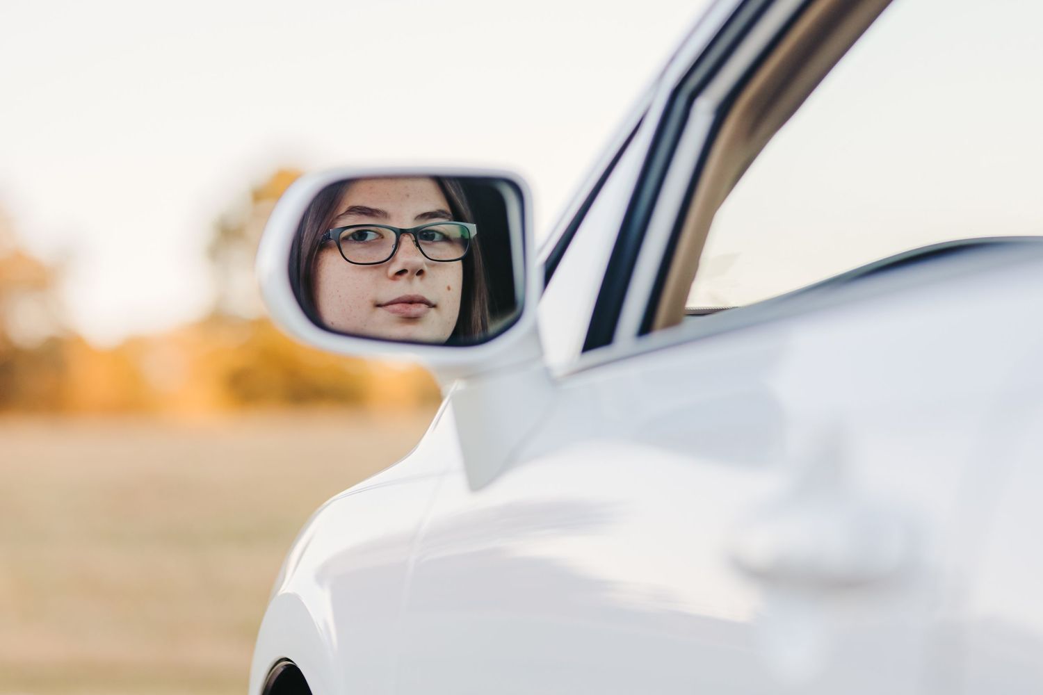 Side mirror reflection on white car during golden hour showing driver wearing glasses.