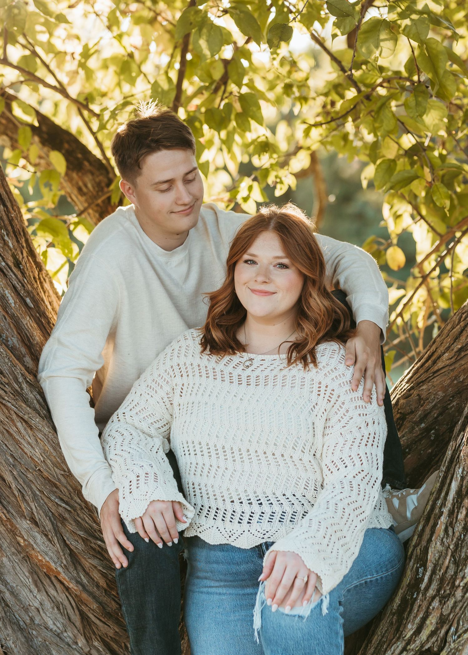 Two people pose together in a tree trunk with golden leaves overhead during sunset.