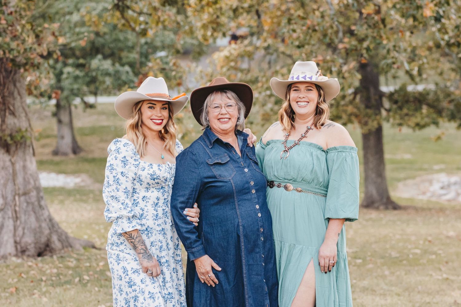 Three people wearing western-inspired outfits pose together in a park setting.