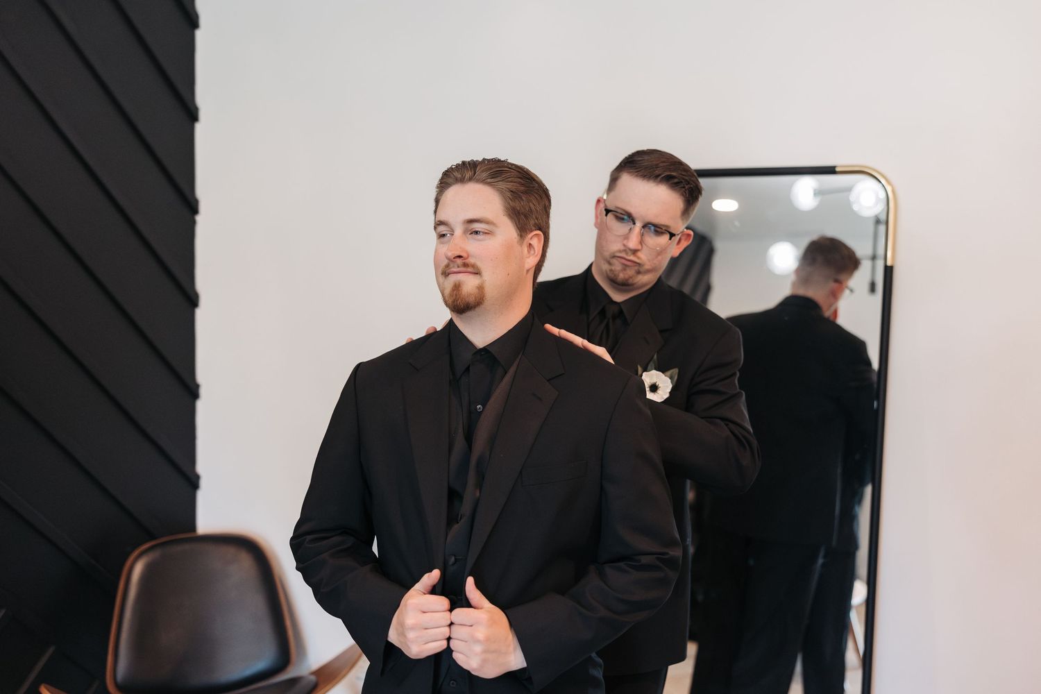 A person in formal black attire adjusts their jacket while standing near a mirror and dark accent wall.