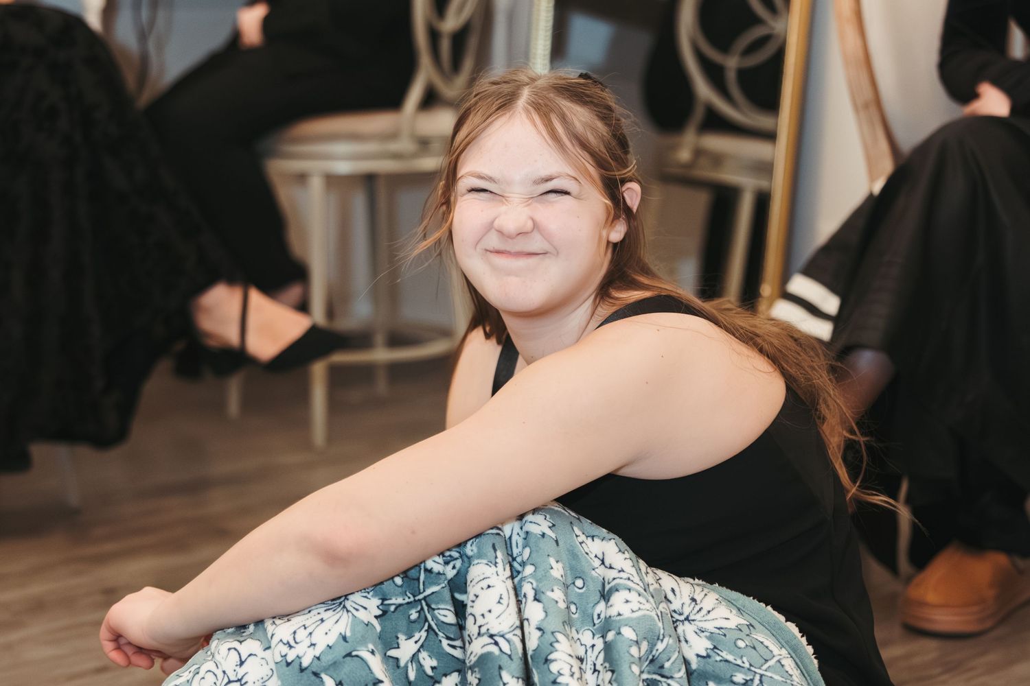 A wedding guest in a black top and patterned skirt sits on the floor at a reception.
