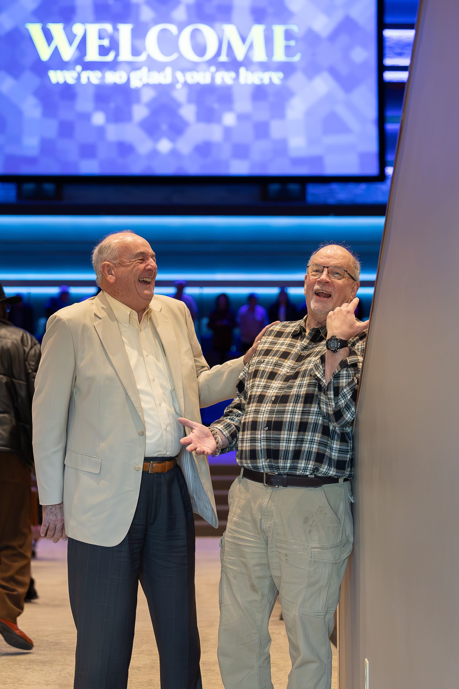 Two men sharing a lighthearted moment under a welcome sign with blue lighting.