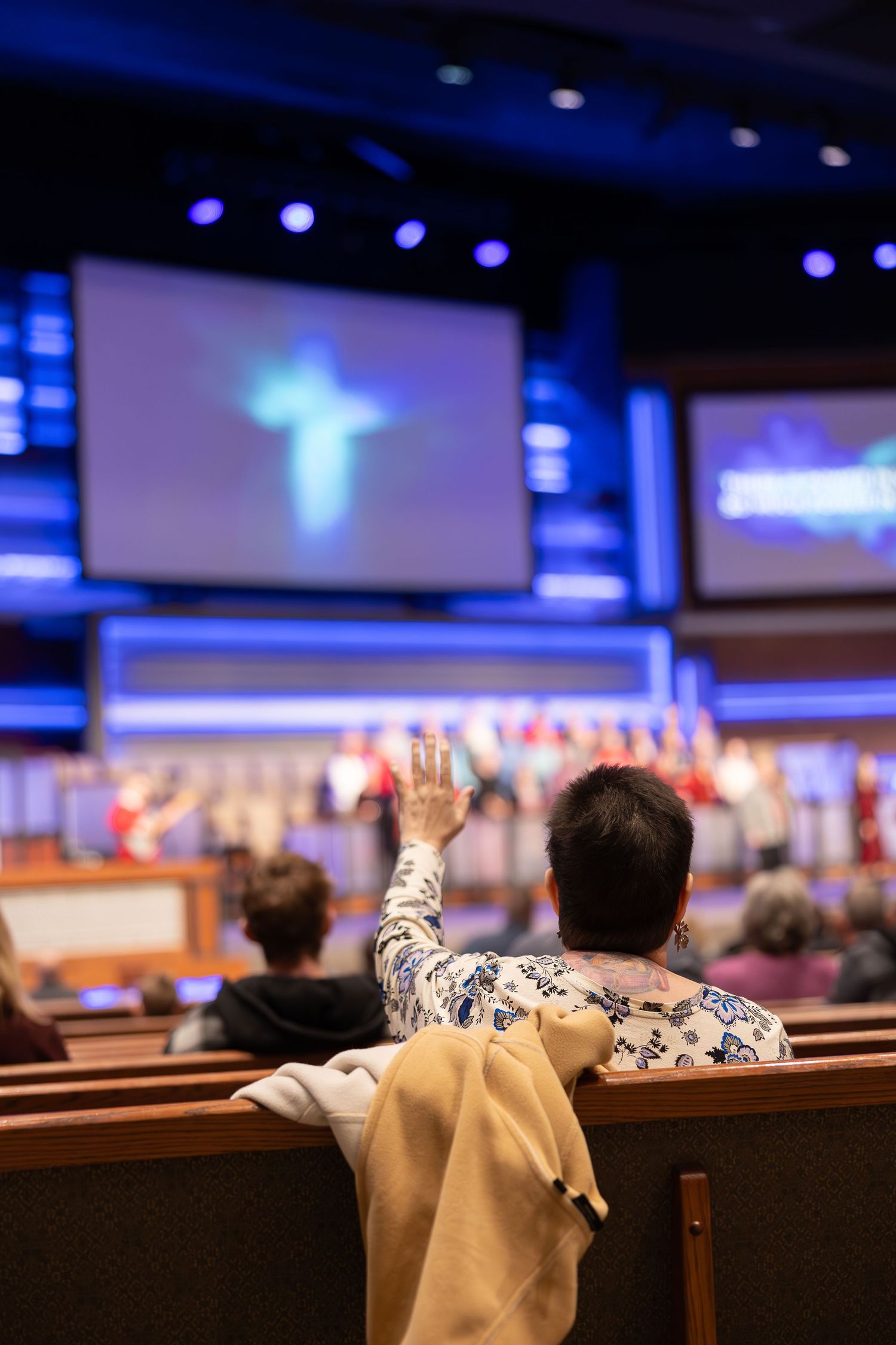 Parishioner raises hand in worship with illuminated cross displayed on screen above.