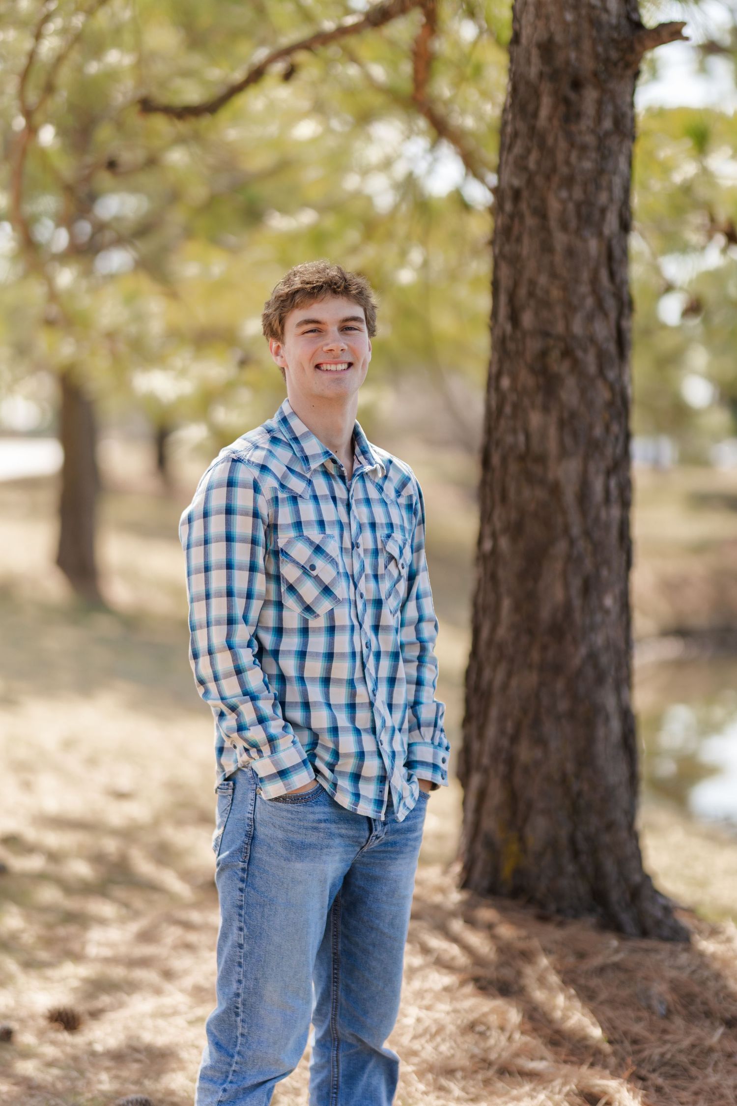 Outdoor portrait of person in blue plaid shirt smiling among pine trees.