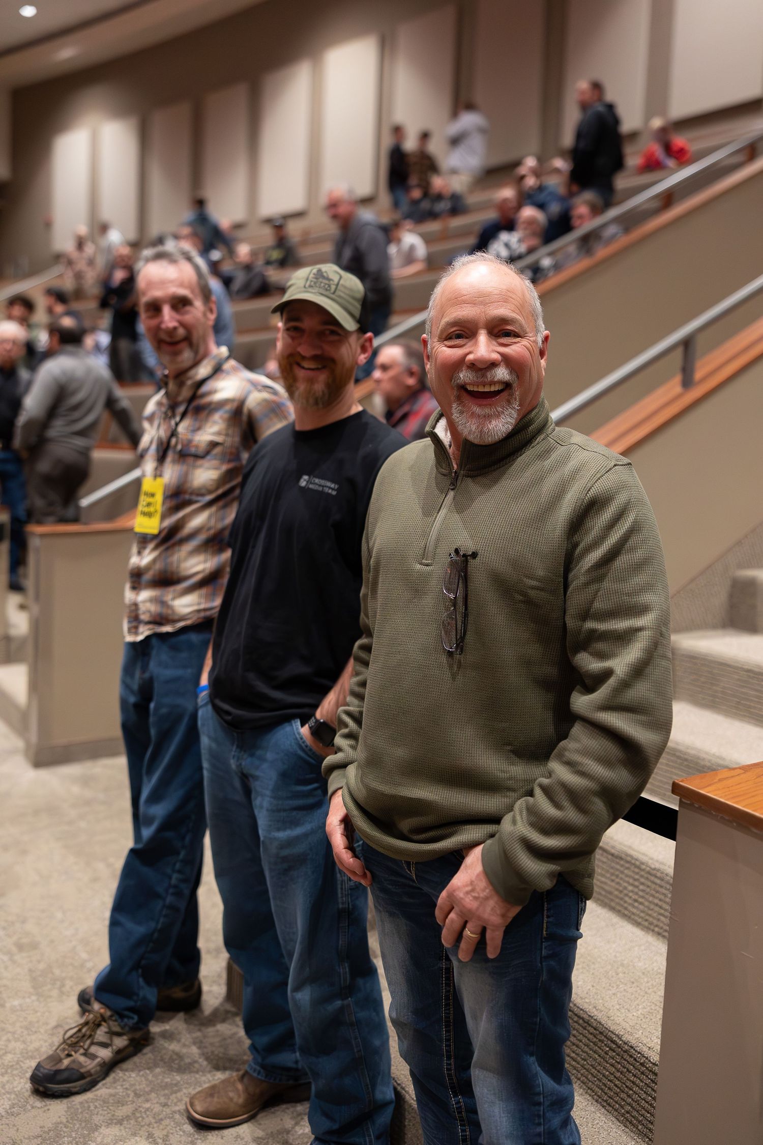 Three people standing together on tiered steps in an auditorium wearing casual clothing and smiling.