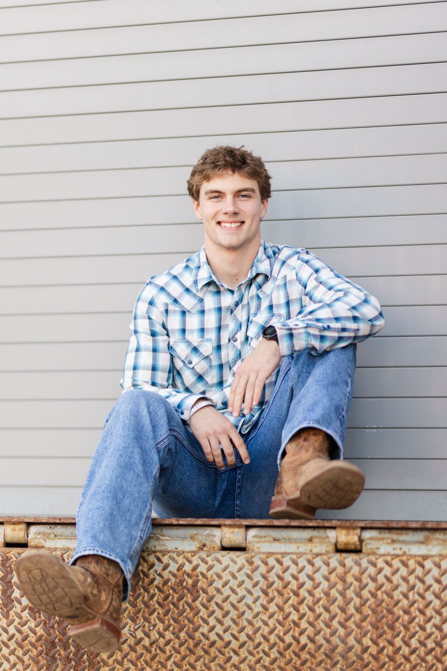 Person in blue plaid shirt and jeans sits casually on metal surface against gray wall background.