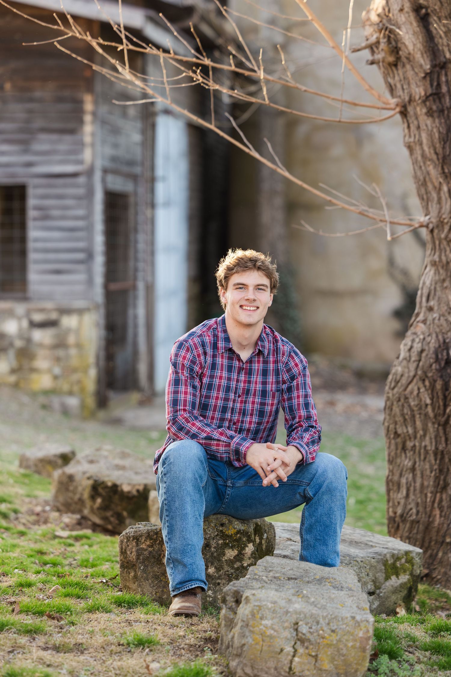 A person in a plaid shirt and jeans sits casually on a large rock with an old wooden building in the background.
