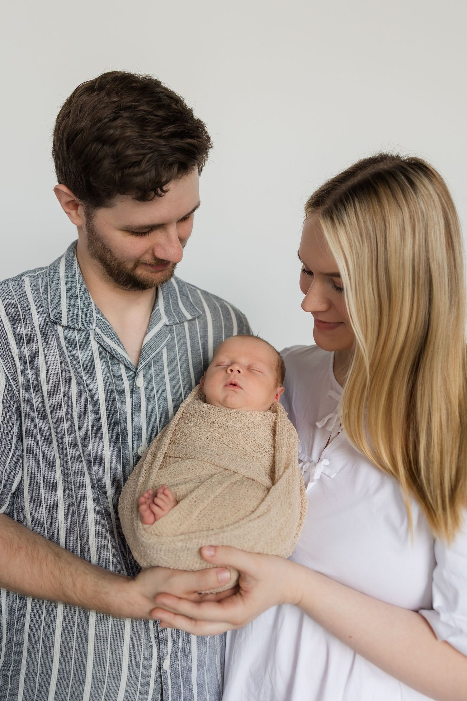 A series of tender moments showing a couple holding their newborn baby wrapped in a beige blanket against a light background.