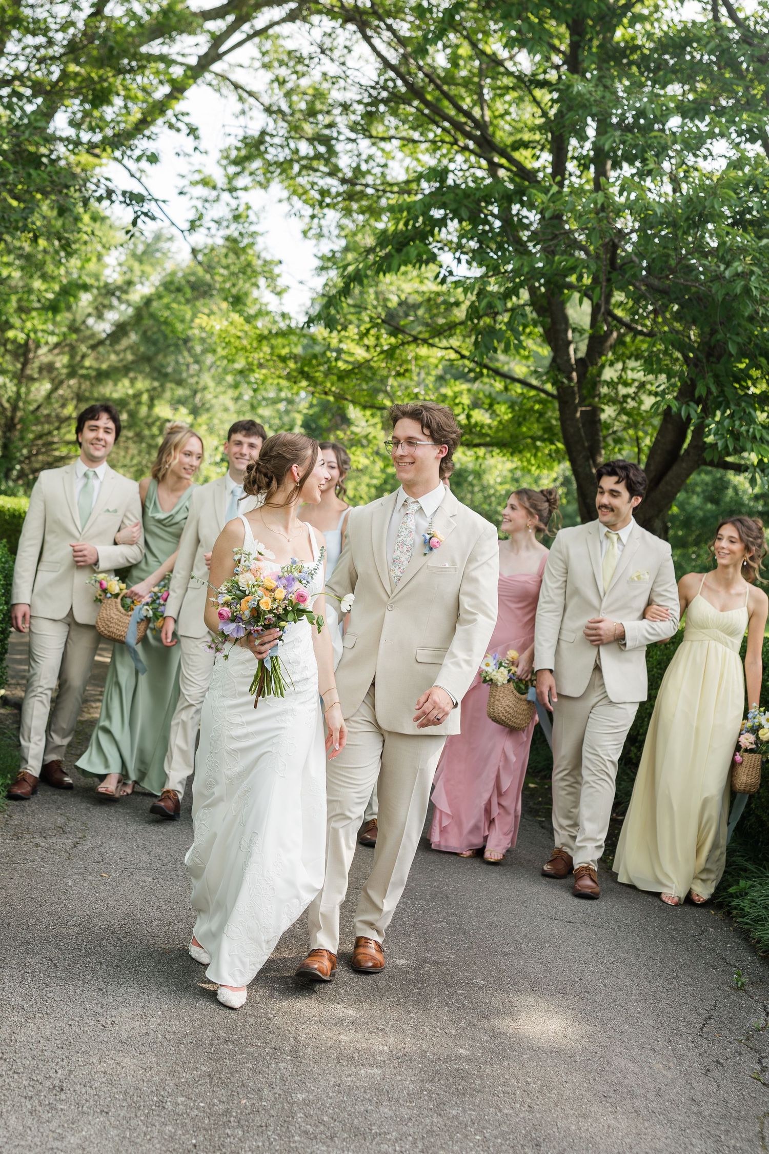 Bride and groom walk with wedding party in sage and blush dresses and tan suits along a tree-lined path.